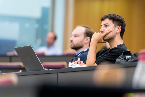 Two men sit in a lecture hall, focused on their laptops during a presentation.