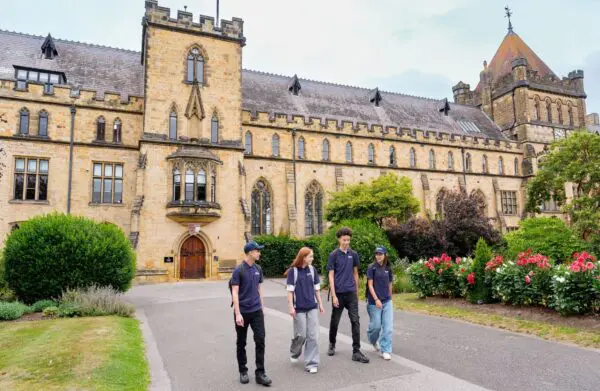 Four individuals walking along a paved path in front of a large historic building with Gothic-style architecture. The building features tall arched windows, decorative stonework, and a tower with battlements. Surrounding the path are manicured gardens with green shrubs and vibrant flowers.