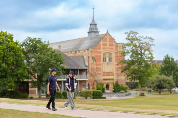 Two people walking along a paved path on a grassy campus with trees and a large historic brick building featuring a tower and cross in the background. The sky is partly cloudy.