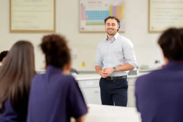 Person standing at the front of a classroom, wearing a light-colored shirt and dark trousers, speaking to a group of seated students. The classroom has science posters on the walls, including a periodic table, and lab-style counters.