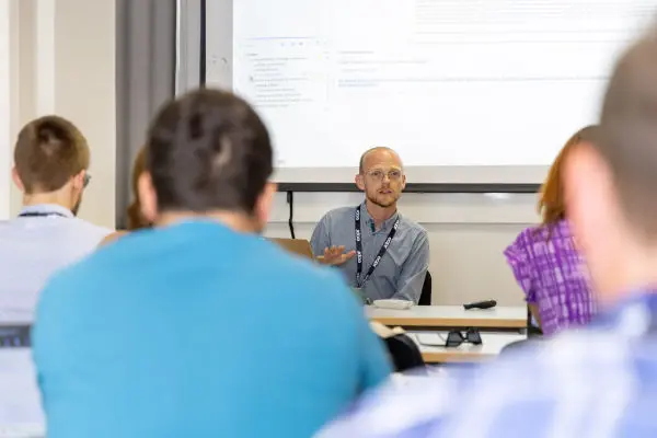 A presenter in a blue shirt gestures while addressing an audience in a classroom setting. Projector screen behind him.