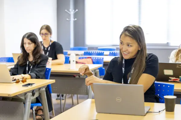 Three women in a classroom setting, each working on a laptop. The woman on the right is gesturing as she talks.
