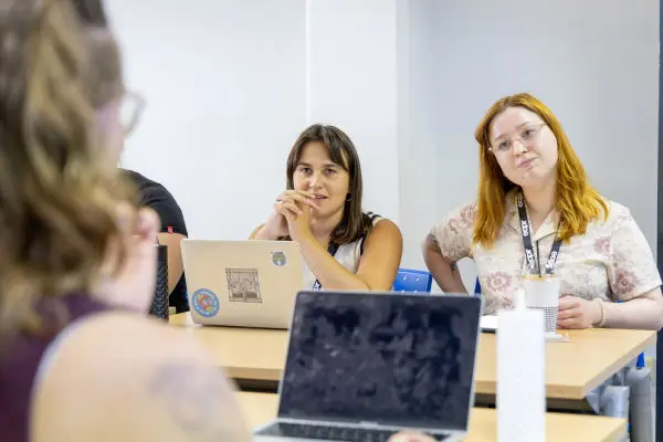 Three people at tables with laptops, two women in focus looking towards a speaker. One has brown hair, the other red.
