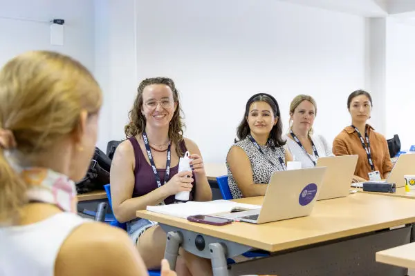 Women in a classroom setting, some smiling and some looking thoughtful, with laptops and ID lanyards. One woman holds a water bottle.
