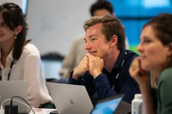 A man wearing a dark shirt and ECPR lanyard smiles with hands clasped under chin. He is seated at a table with laptops, in a classroom setting with other participants visible in the foreground and background.