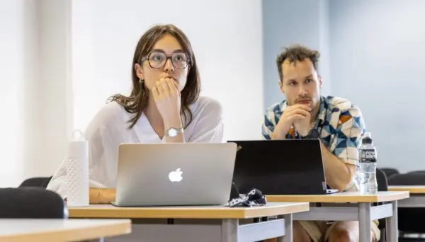 Two students in a classroom, a woman with glasses in the foreground and a man behind her, both looking intently at their laptops.