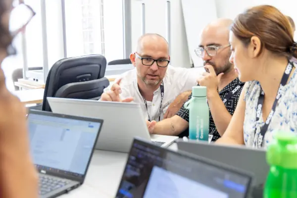 Three colleagues in a bright office, gathered around a laptop, engaged in discussion and collaborative work.