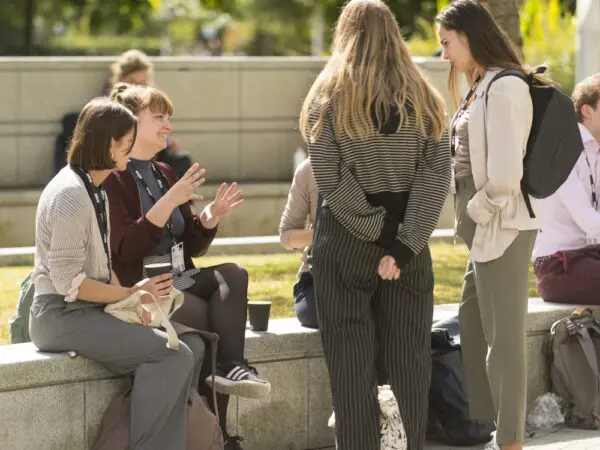 Four people are casually conversing outdoors on a sunny day, seated and standing near a stone ledge.
